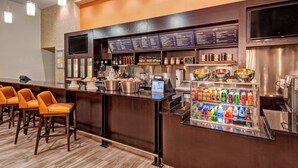 A hotel bar area with a long counter, orange bar stools, a beverage cooler, and a menu board.
