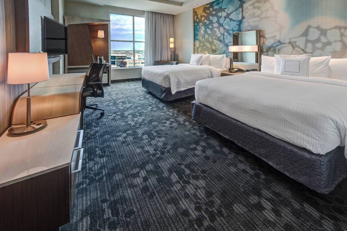 A hotel room with two beds, a desk, a chair, a TV, and a large window offering a city view.