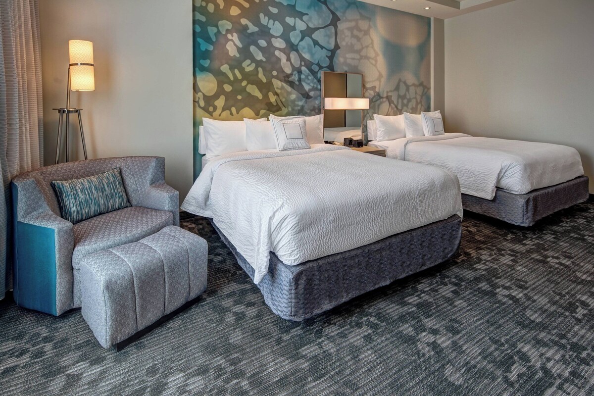 A hotel room with two beds, a sofa, a standing lamp, and a large abstract wall mural.