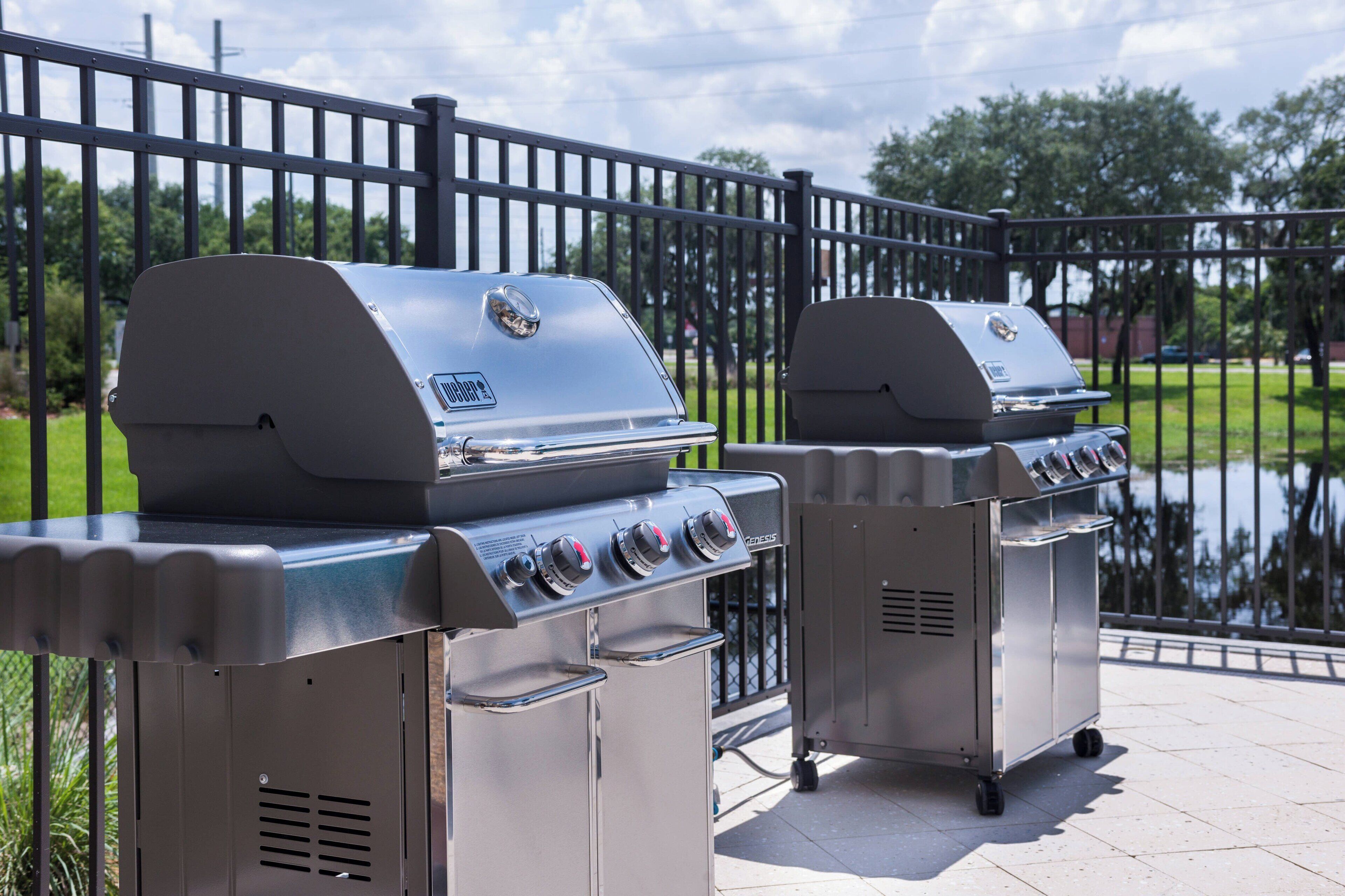 Two portable gas grills on a patio with a metal fence and a body of water in the background.