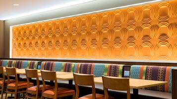 Un ristorante con tavoli e sedie in legno, una zona salotto con una panca colorata e un muro decorato con un motivo circolare.