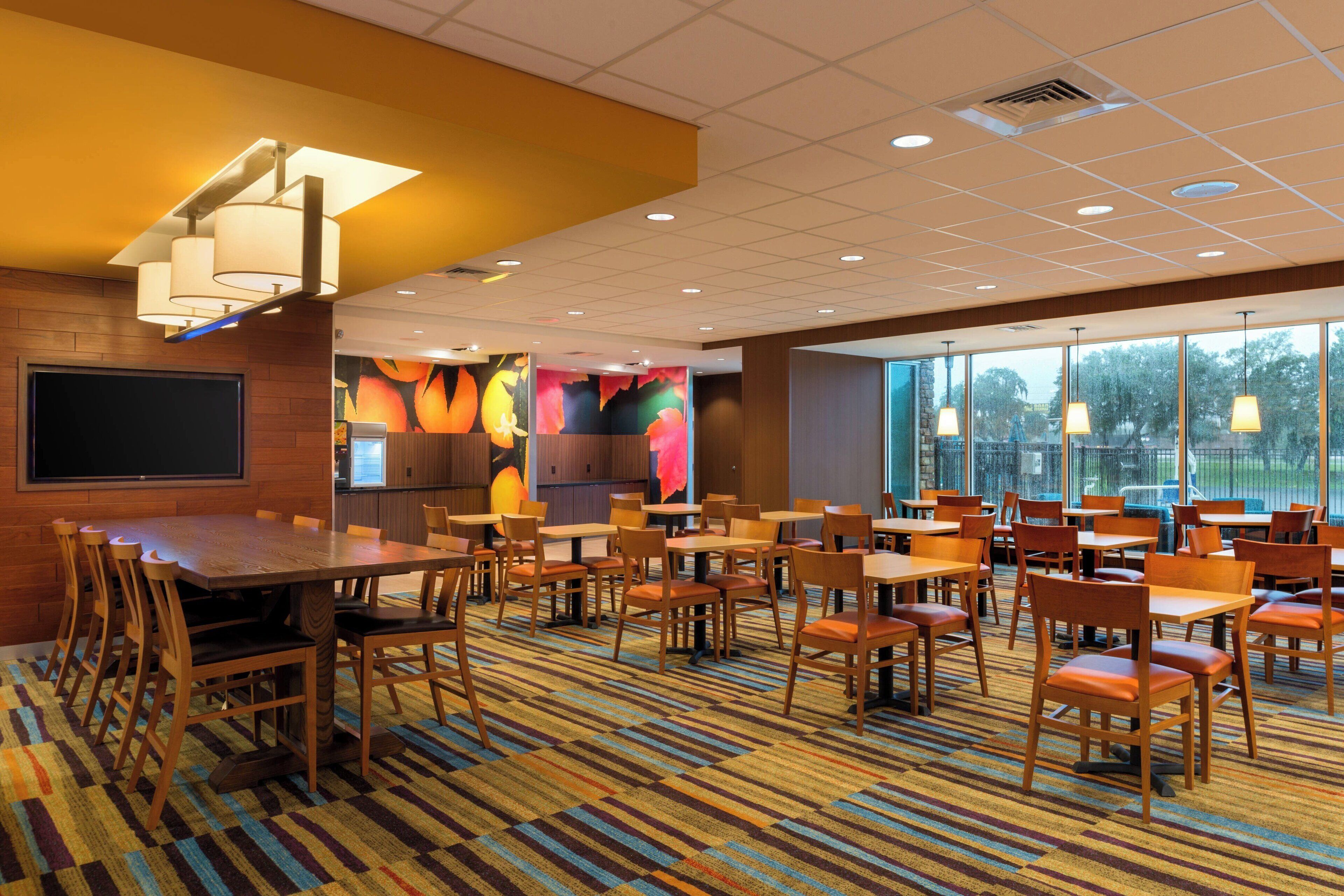 A hotel dining area with a large wooden table and chairs, a colorful carpet, and large windows offering a view of trees.