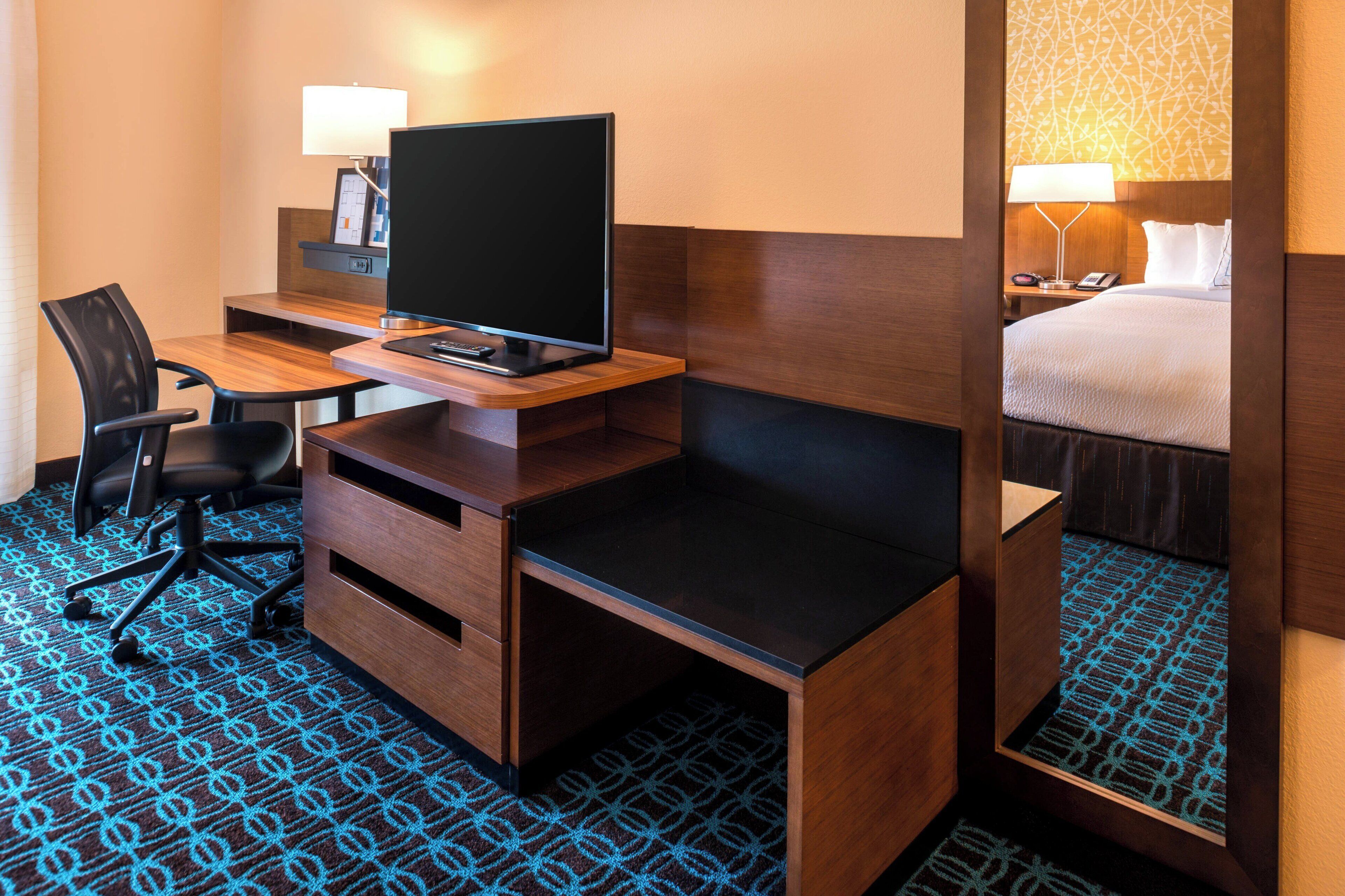 Ein Hotelzimmer mit einem Holztisch, einem Sessel, einem Fernseher, einem Bett und einem Nachttisch mit einer Lampe.