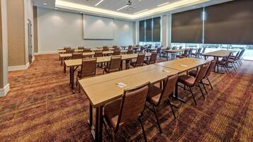 Uma sala de conferências com várias mesas e cadeiras, piso coberto de carpete, grandes janelas e um projetor.