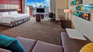 Habitación de hotel con cama, escritorio y zona de estar, ofreciendo vistas a la ciudad a través de grandes ventanales.