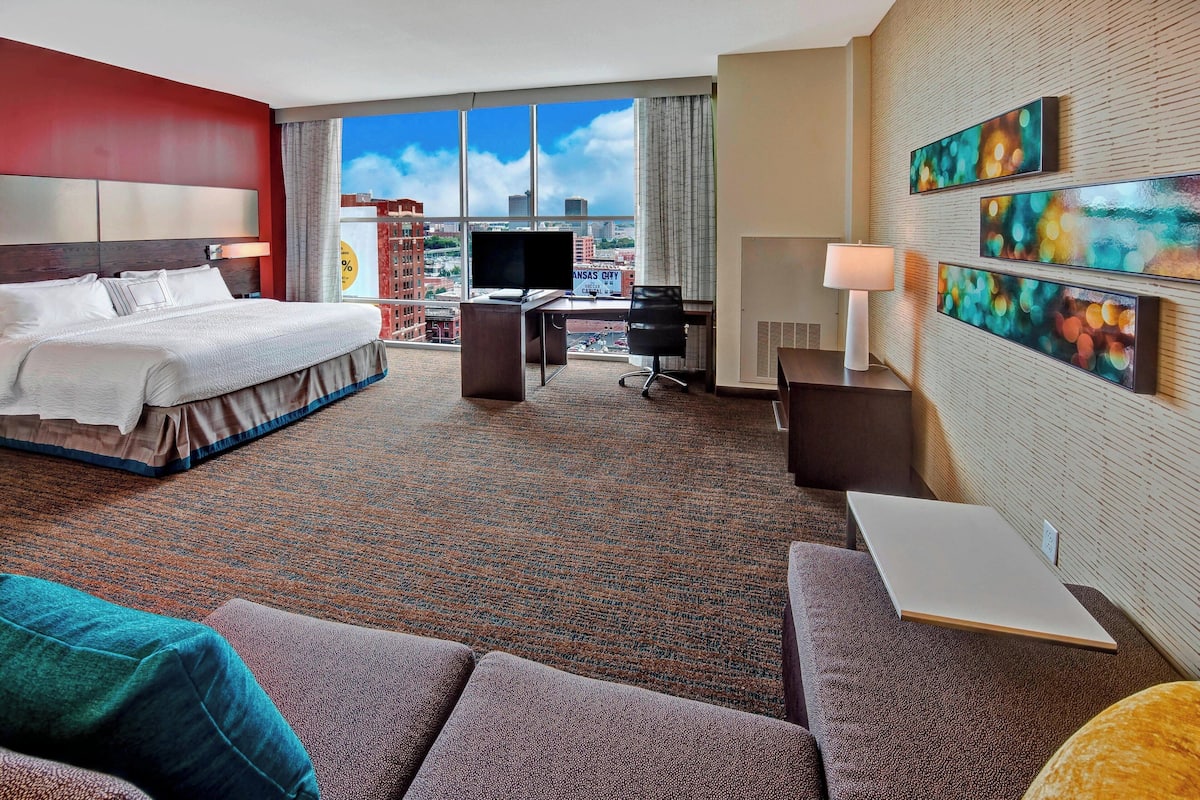 Habitación de hotel con cama, escritorio y zona de estar, ofreciendo vistas a la ciudad a través de grandes ventanales.