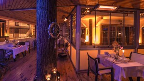 Un comedor cálidamente iluminado con suelos de madera y mesas dispuestas para comer, un llamativo centro de mesa con un tronco de árbol grande, y vistas al exterior a través de amplios ventanales.