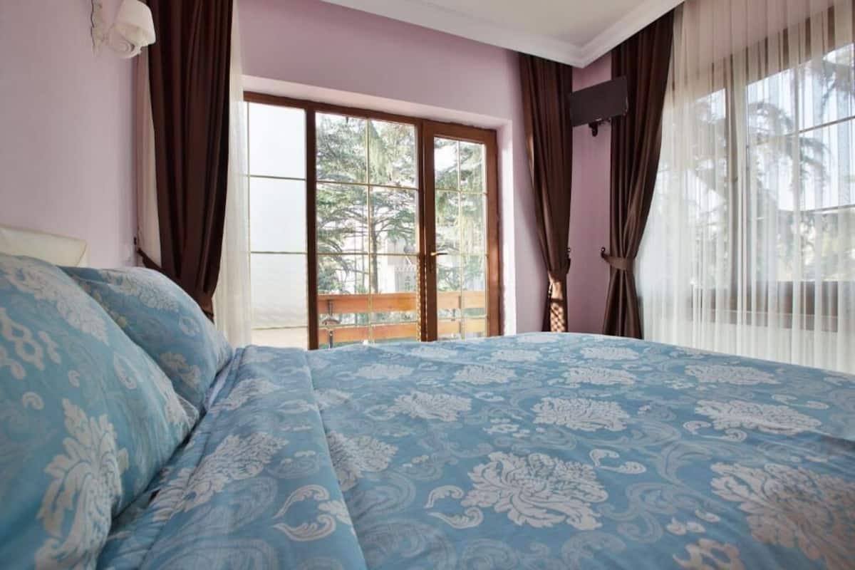 Ein Schlafzimmer mit einem großen Bett, einem Fenster mit Vorhängen und Blick ins Freie.