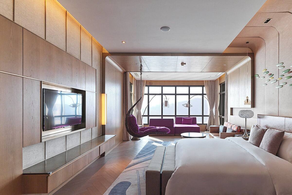 Une chambre d’hôtel moderne dotée d’un grand lit, d’une télévision, d’un coin salon avec un canapé et des fauteuils, et d’une grande fenêtre offrant une vue dégagée.