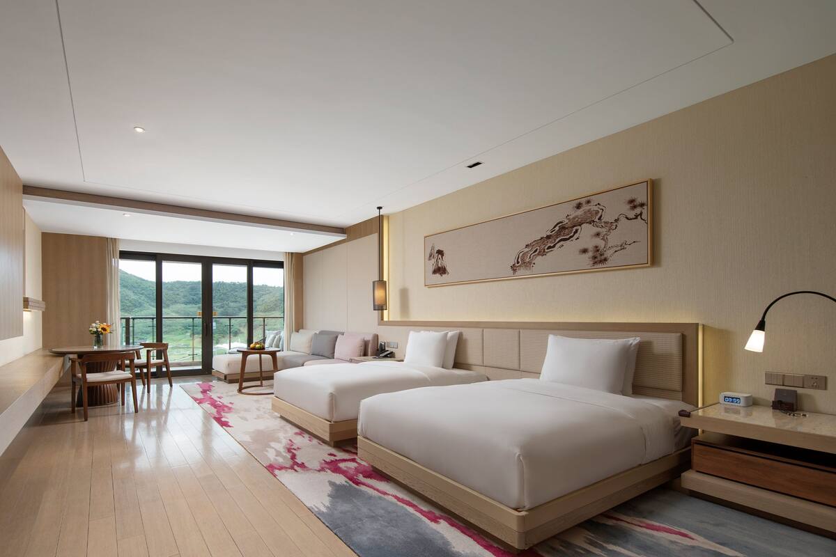 Une chambre d’hôtel dotée d’une grande télévision à écran plat, de deux lits, d’un bureau avec des chaises, d’un balcon offrant une vue sur les montagnes et d’un tableau accroché au mur.