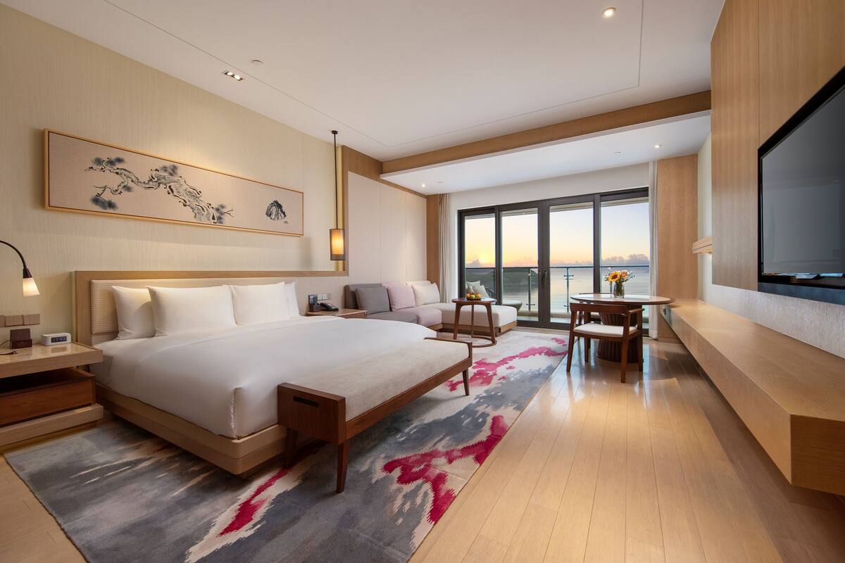 Une chambre d’hôtel moderne, dotée d’un grand lit, d’un bureau, d’une chaise et offrant une vue sur le coucher de soleil par la fenêtre.