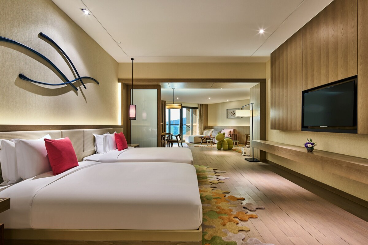 A hotel room with two beds, a TV, a wooden wall panel, and a view of the city.