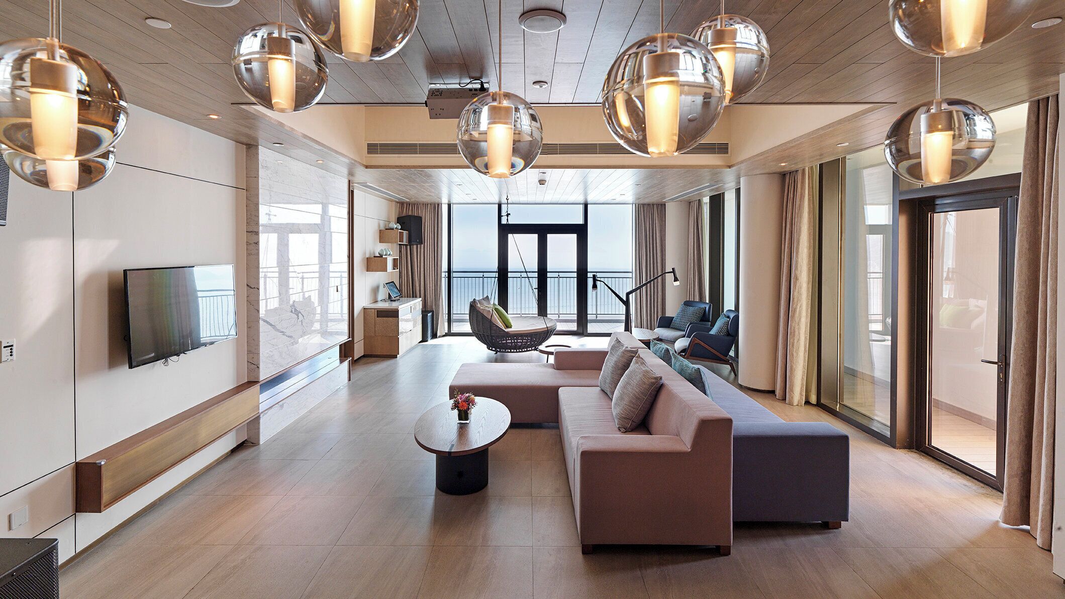 Una sala de estar moderna con un sofá, una mesa de centro y un sillón colgante. Hay un televisor montado en la pared y un ventanal que ofrece vistas al exterior.