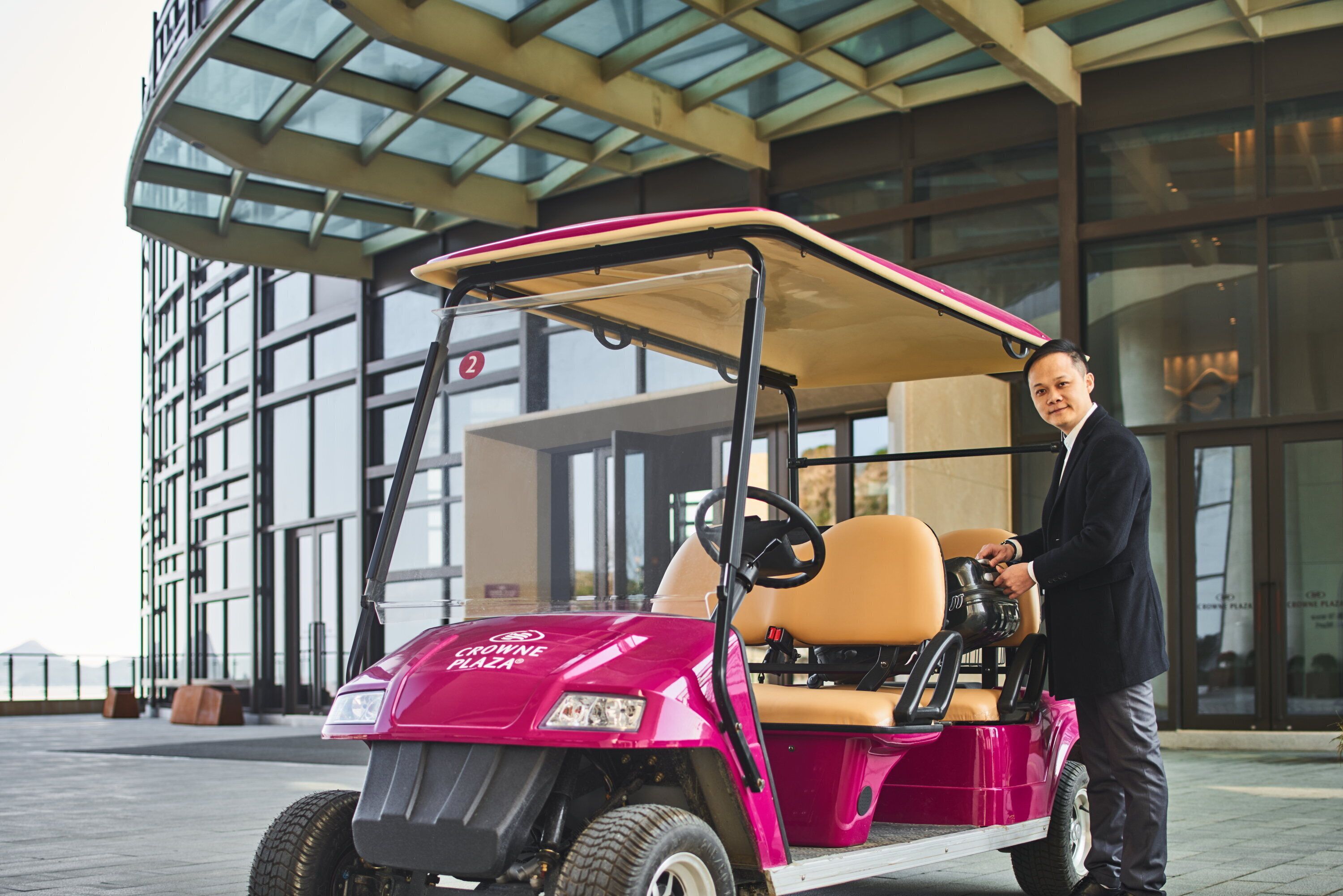 A golf cart with a driver, parked in front of a modern building with large windows.