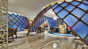 Un intérieur moderne doté d’un design original en forme d’arc, comprenant un espace de détente central avec des tables et des chaises, ainsi qu’un mur décoratif à motifs texturés.