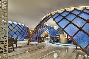 Un intérieur moderne doté d’un design original en forme d’arc, comprenant un espace de détente central avec des tables et des chaises, ainsi qu’un mur décoratif à motifs texturés.