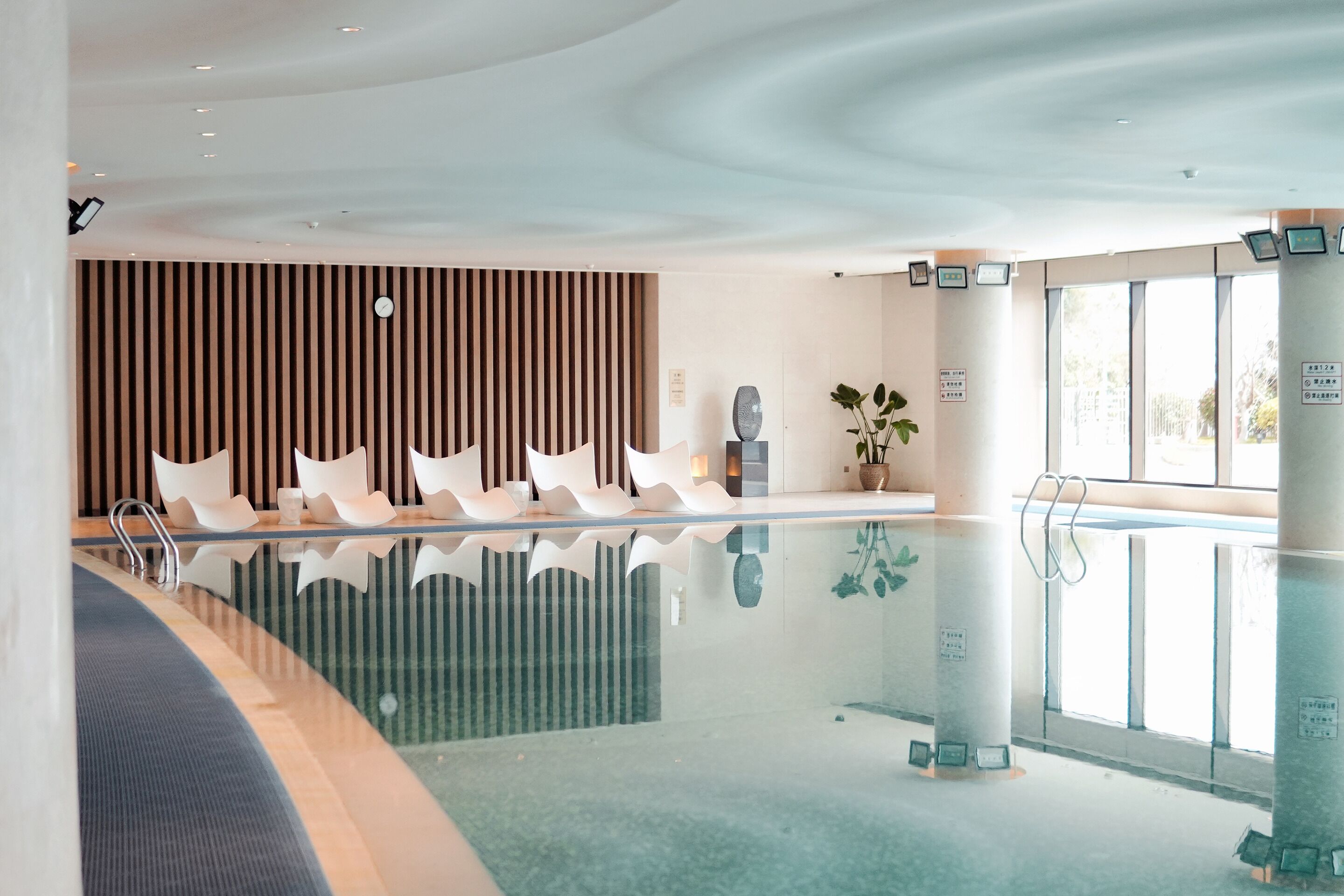 Un área de piscina interior moderna con piso reflectante, asientos a lo largo de la piscina y una pared de listones de madera al fondo.