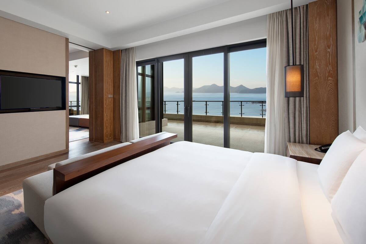 Une chambre d’hôtel dotée d’un grand lit, d’un téléviseur à écran plat fixé au mur, d’un balcon offrant une vue sur la mer et les montagnes, et d’une table de chevet en bois avec une lampe.