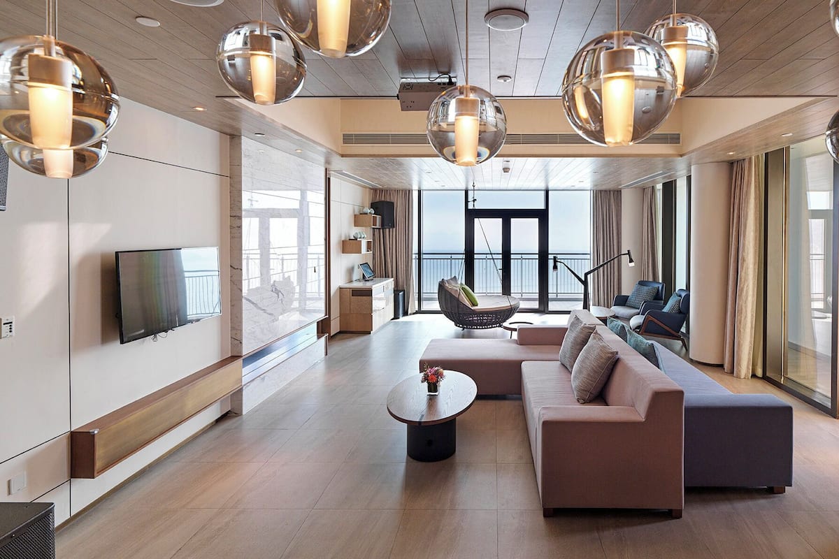Un salon moderne avec un canapé, une table basse et un fauteuil suspendu. Un téléviseur est fixé au mur et une grande fenêtre offre une vue sur l’extérieur.