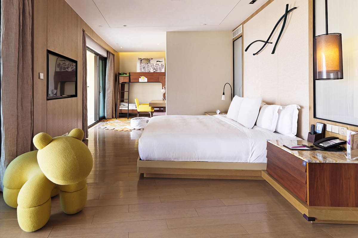 Une chambre d’hôtel avec un grand lit, un bureau, une chaise, un ours en peluche et une grande fenêtre.