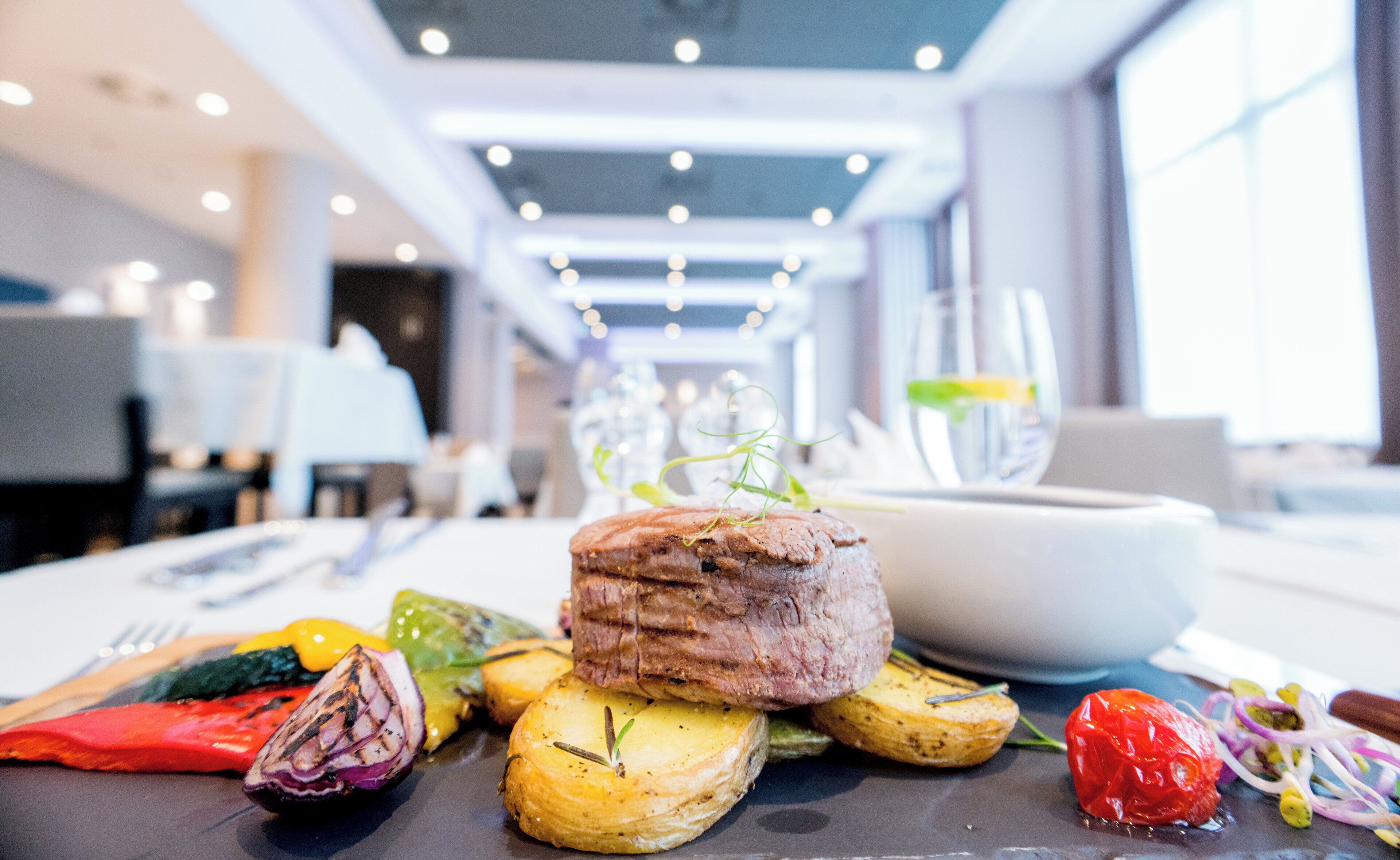 Una mesa de comedor con diversos alimentos, incluyendo un trozo de carne, verduras asadas y un vaso de agua.