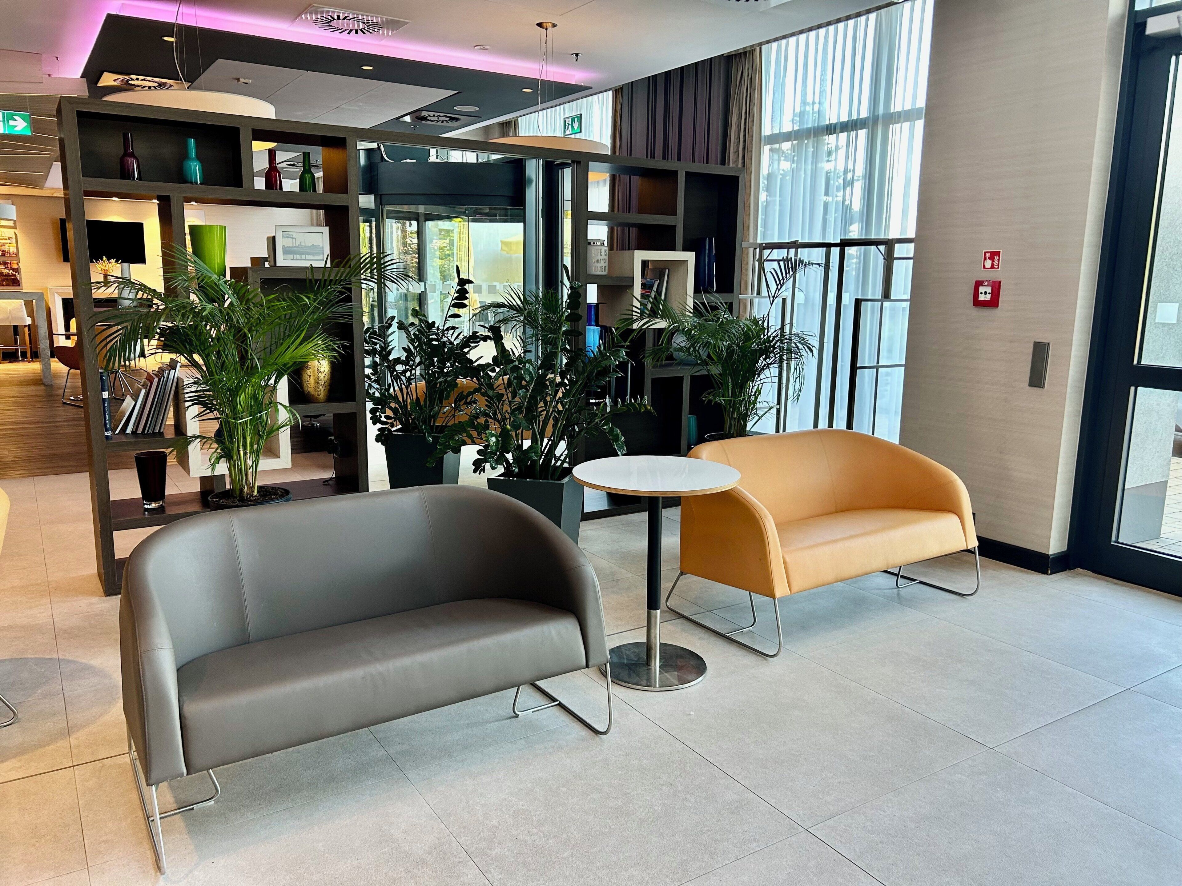 A modern lobby with a grey sofa, an orange armchair, a small round table, and potted plants.