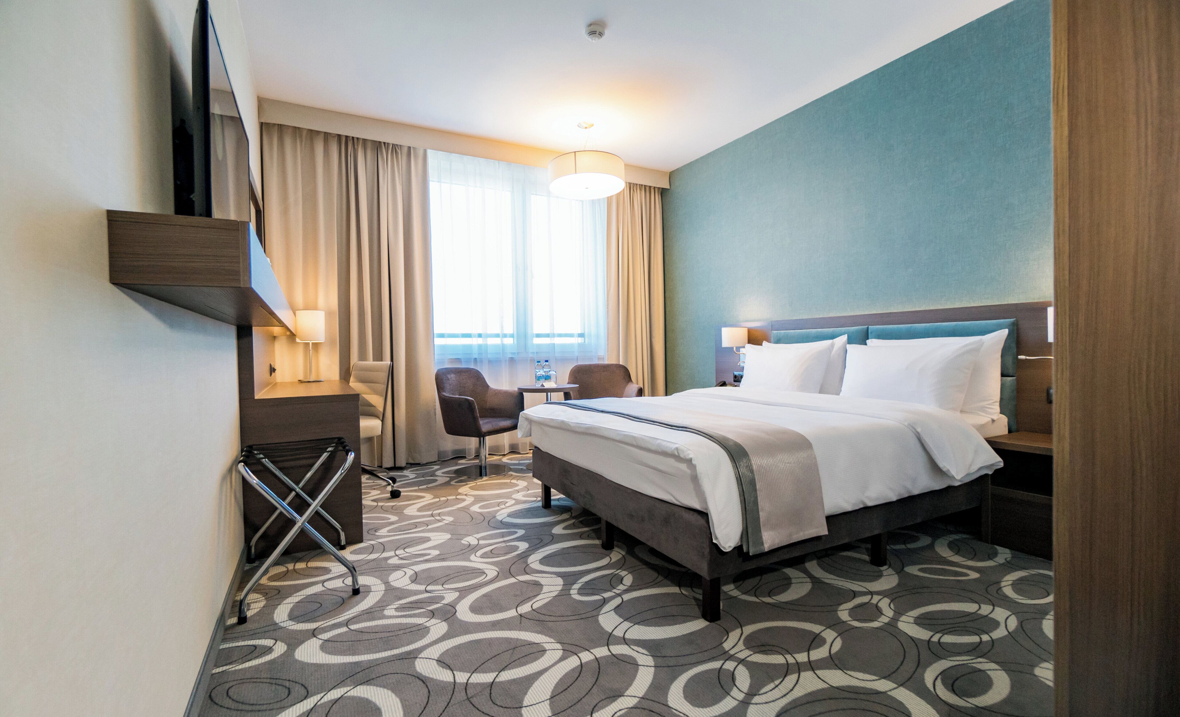 Habitación de hotel con cama, escritorio, silla, lámpara y vistas al exterior.