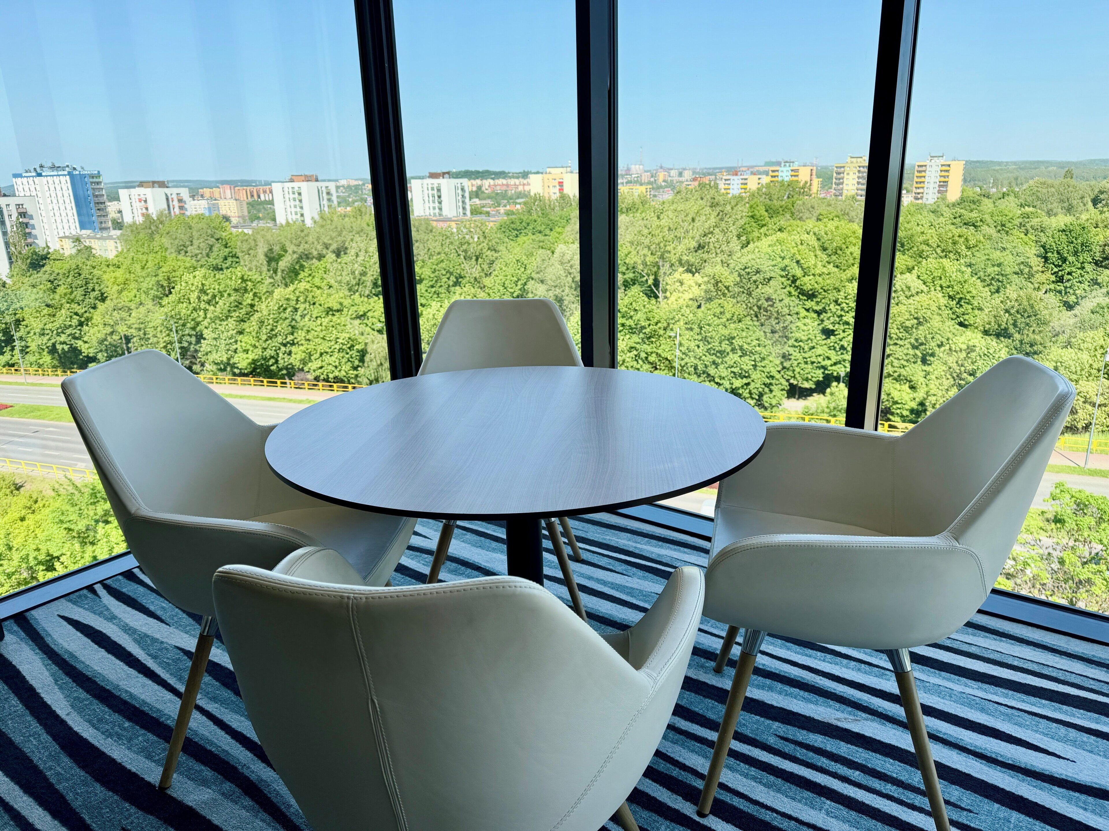 A room with a round table and four chairs, large windows with a cityscape view, and a striped carpet.