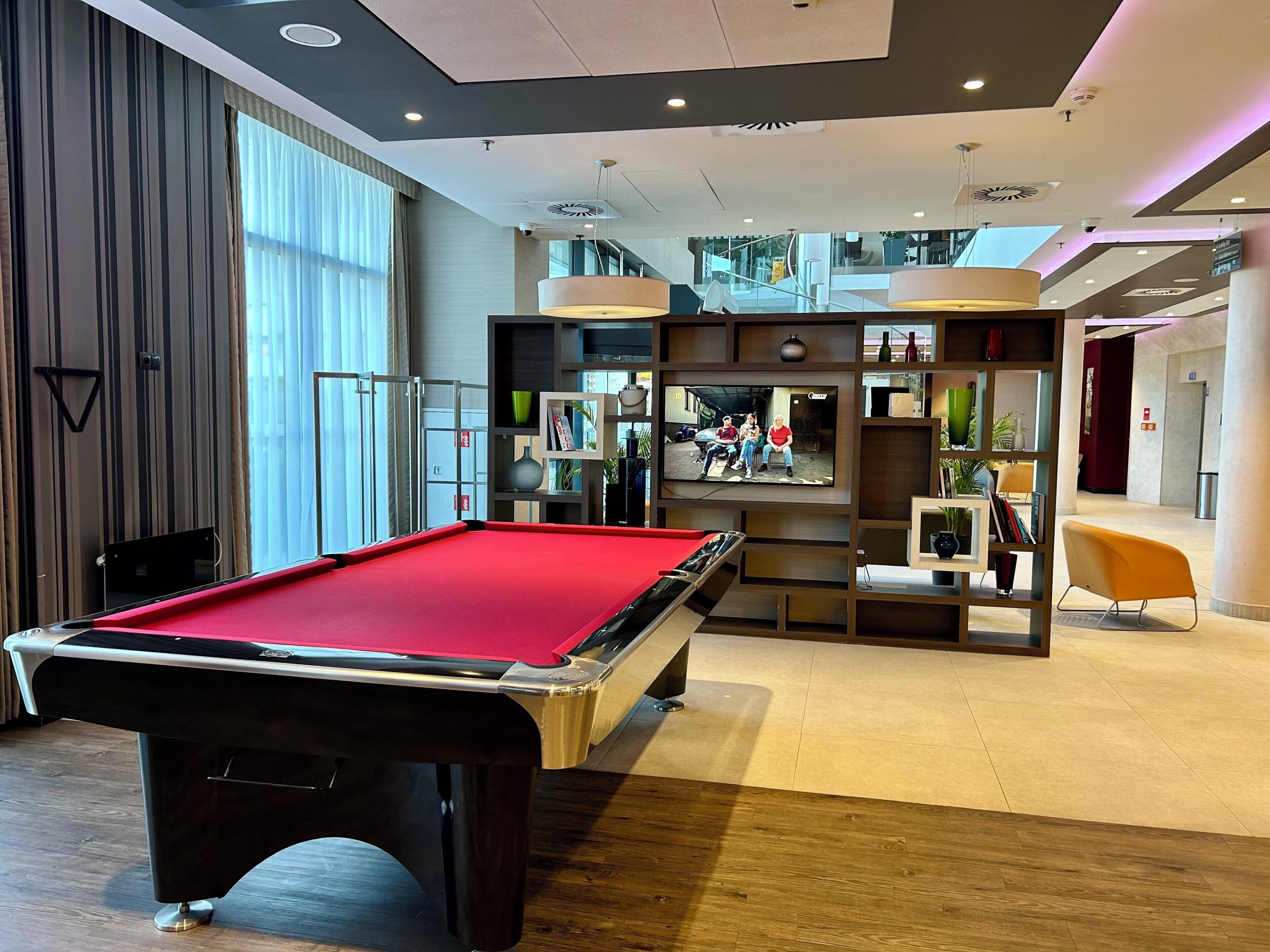 A pool table in a modern hotel lobby with a television displaying a scene, a seating area with a yellow chair, and a bookshelf with various items.