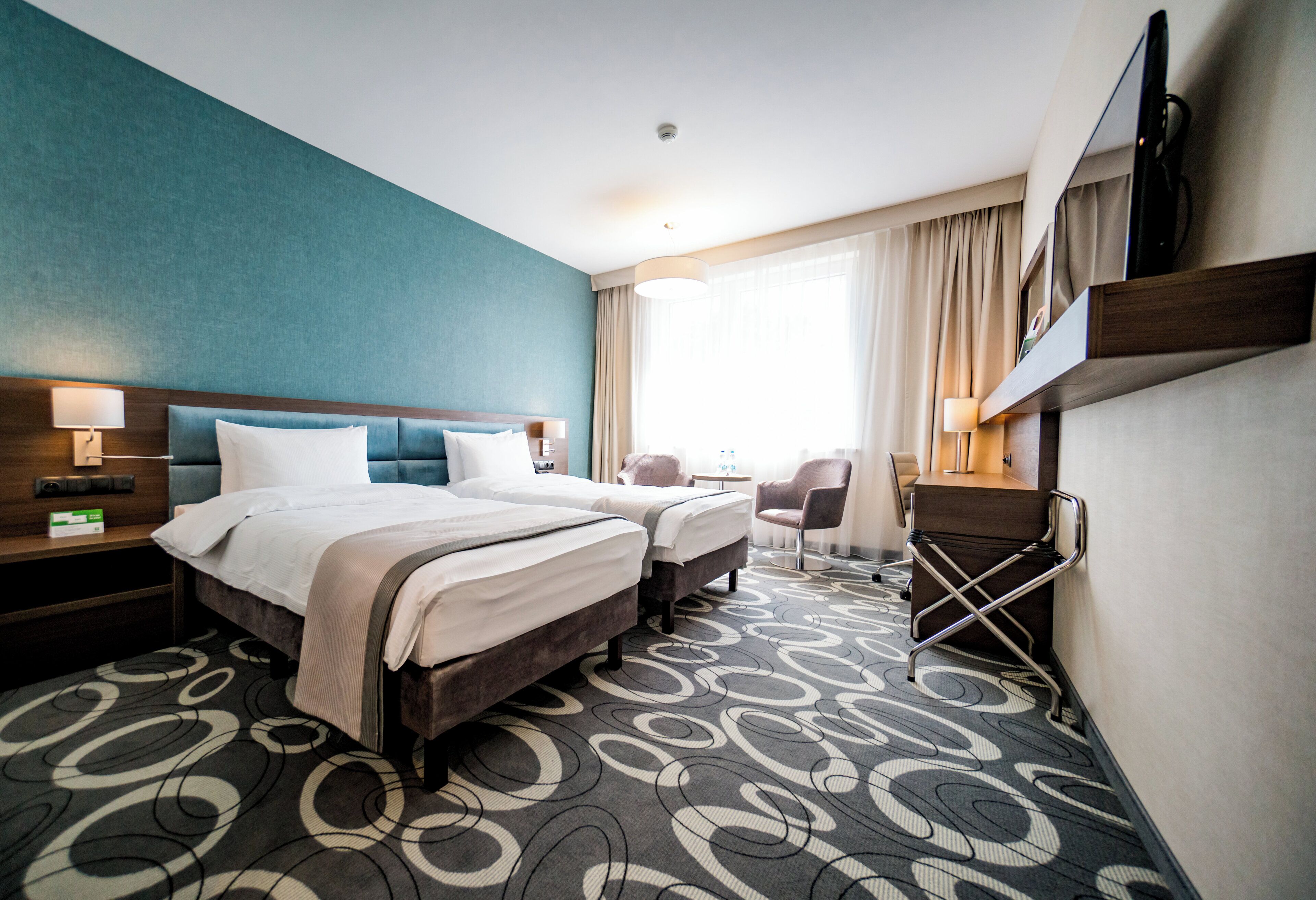 A hotel room with two beds, a desk, a chair, a TV, and a large window with curtains.