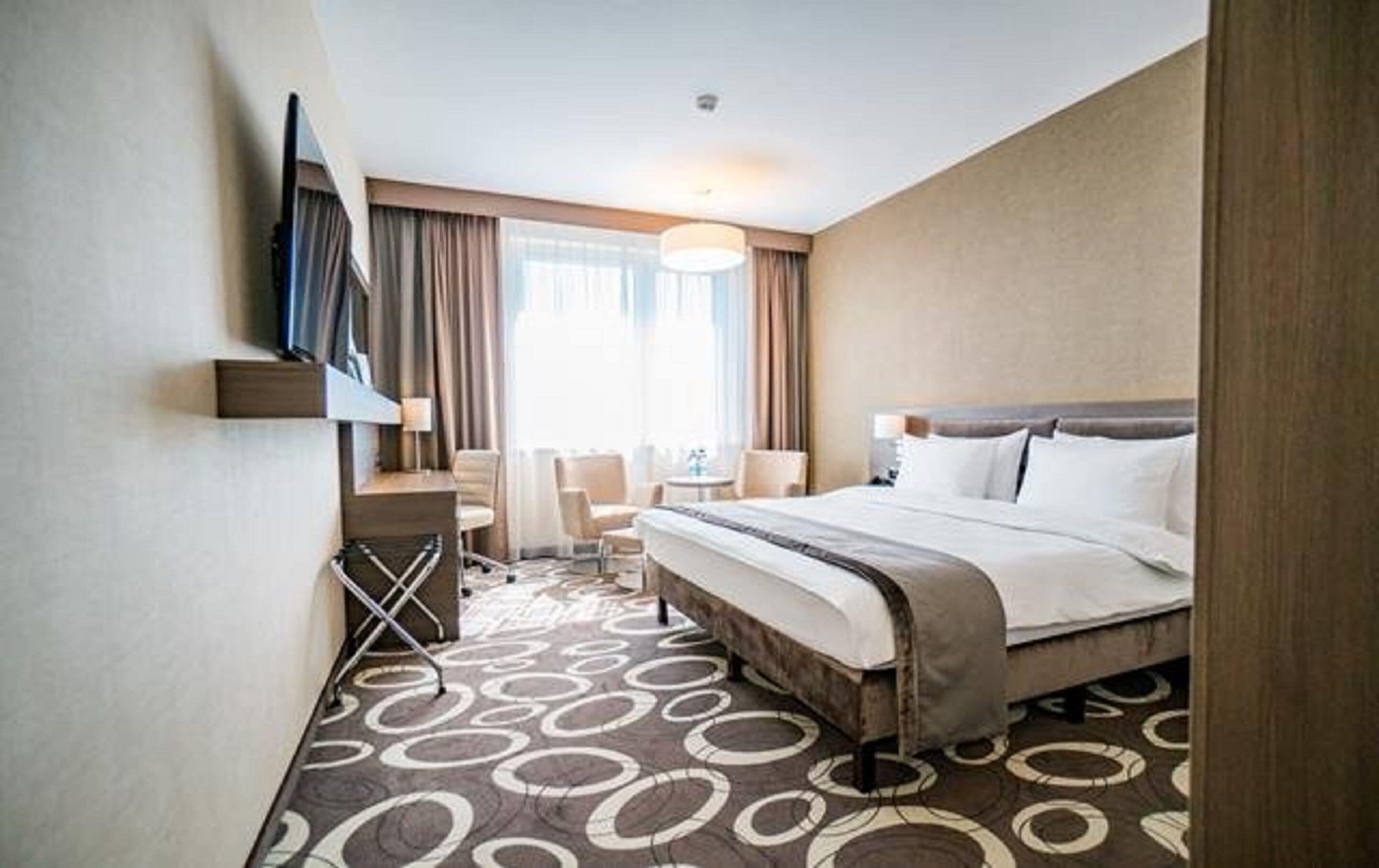 Une chambre d’hôtel avec un grand lit, un bureau avec une chaise, un téléviseur fixé au mur et une fenêtre avec des rideaux.
