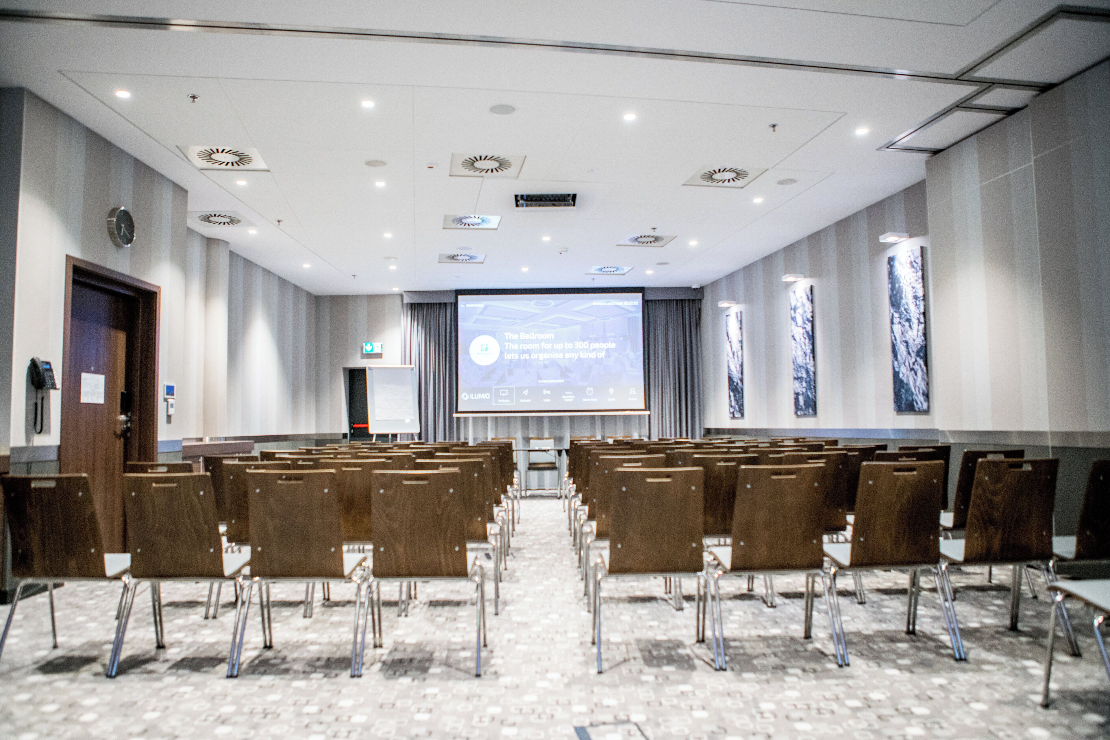 A conference room with rows of chairs, a projection screen, and a clock on the wall.