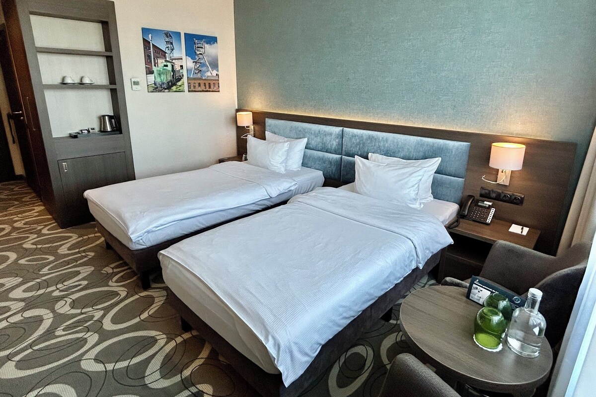 A hotel room with two beds, a small table with a bottle and glass, a nightstand with a phone, and a wall-mounted shelf with a kettle.