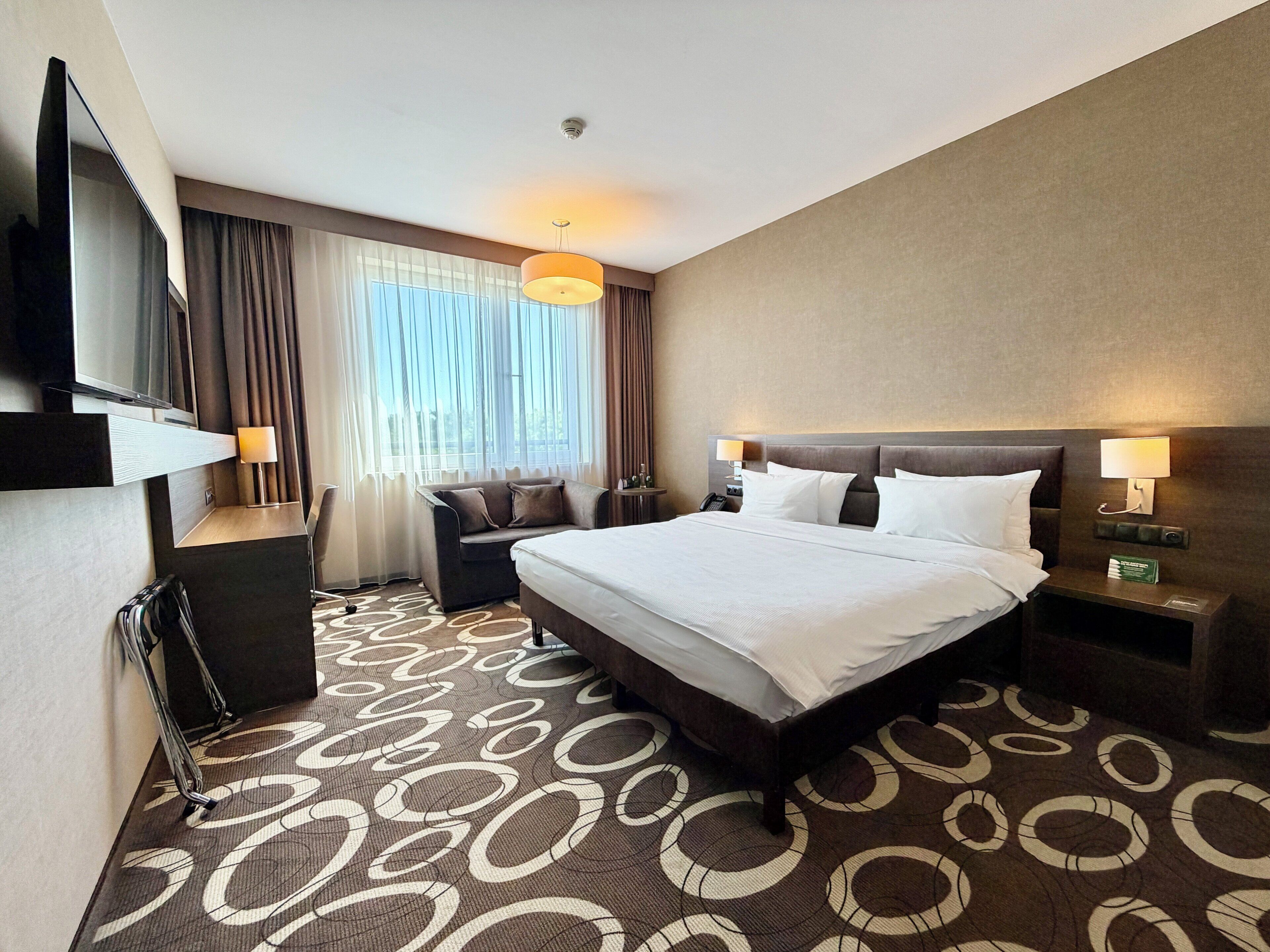 A hotel room with a large bed, a sofa, a TV, and a desk with a lamp.