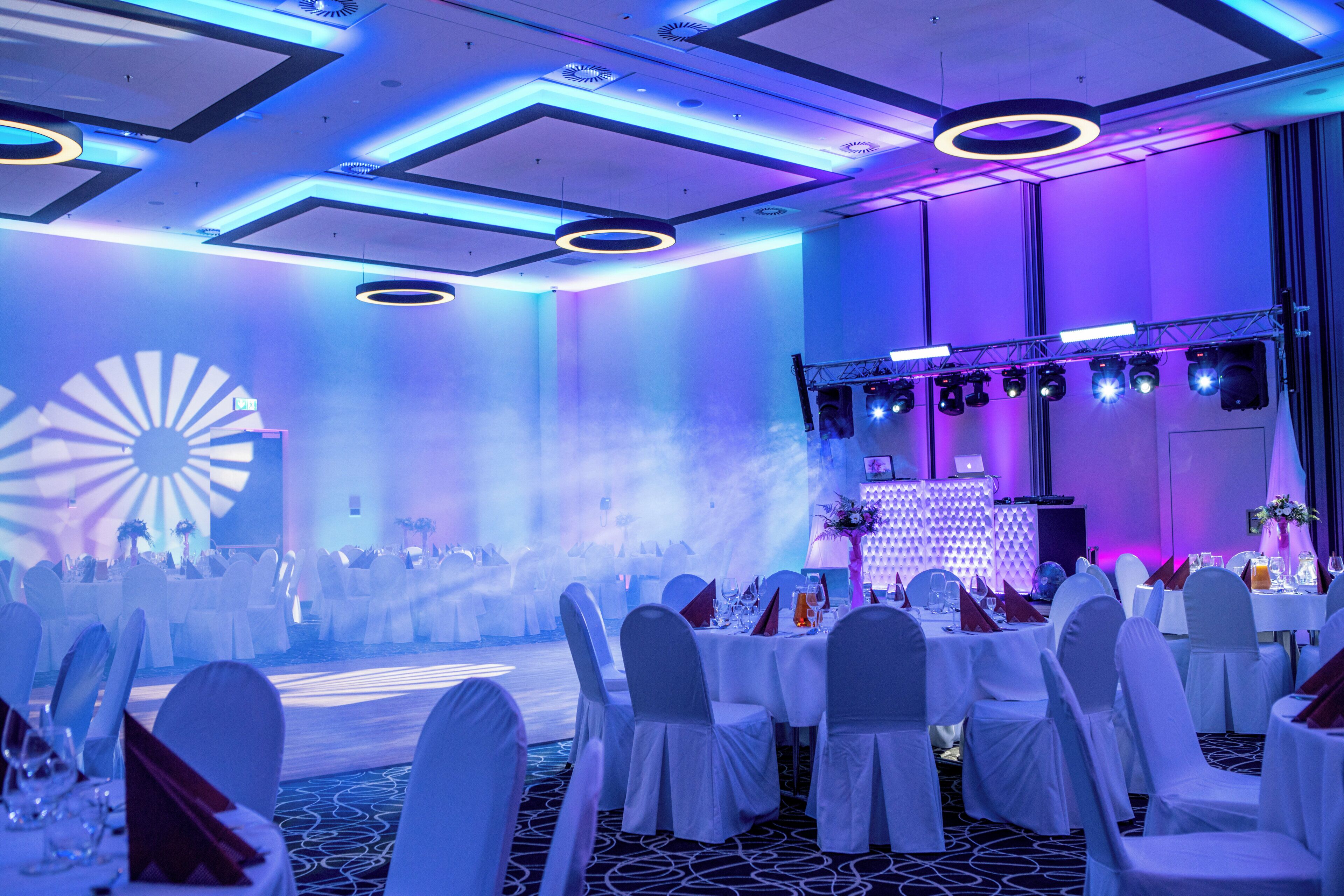 Un salón de banquetes con mesas redondas, sillas blancas y un escenario con equipo de iluminación.