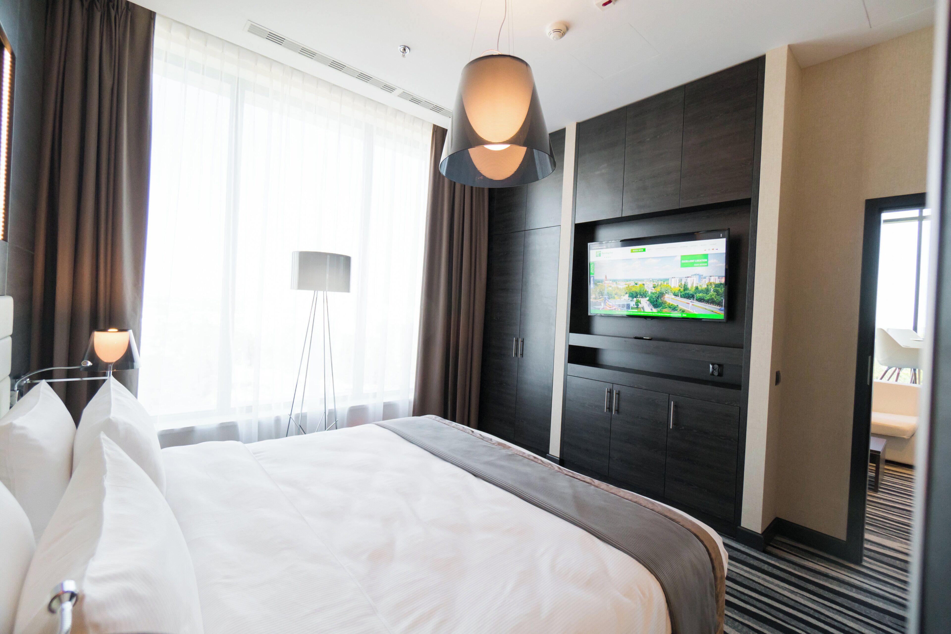 Habitación de hotel con una cama grande, un televisor de pantalla plana en la pared, un escritorio con una lámpara y vistas al exterior por la ventana.