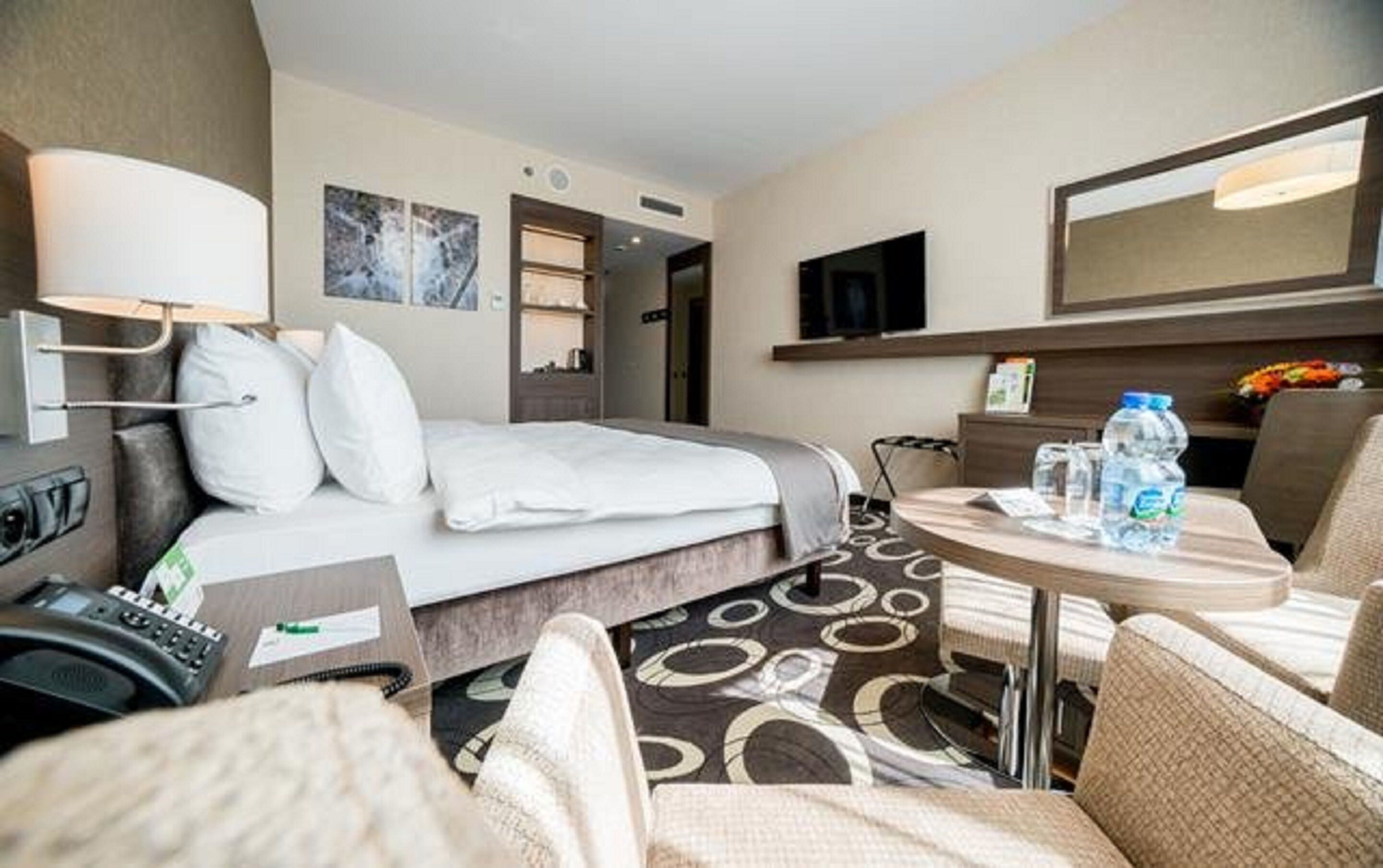 A hotel room with a bed, a desk, a chair, a table, a TV, and a bathroom visible through an open door.