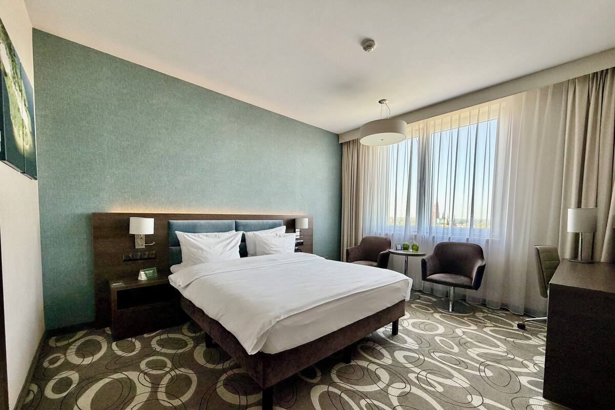 A hotel room with a bed, a desk, two chairs, a small table, and a window with curtains.