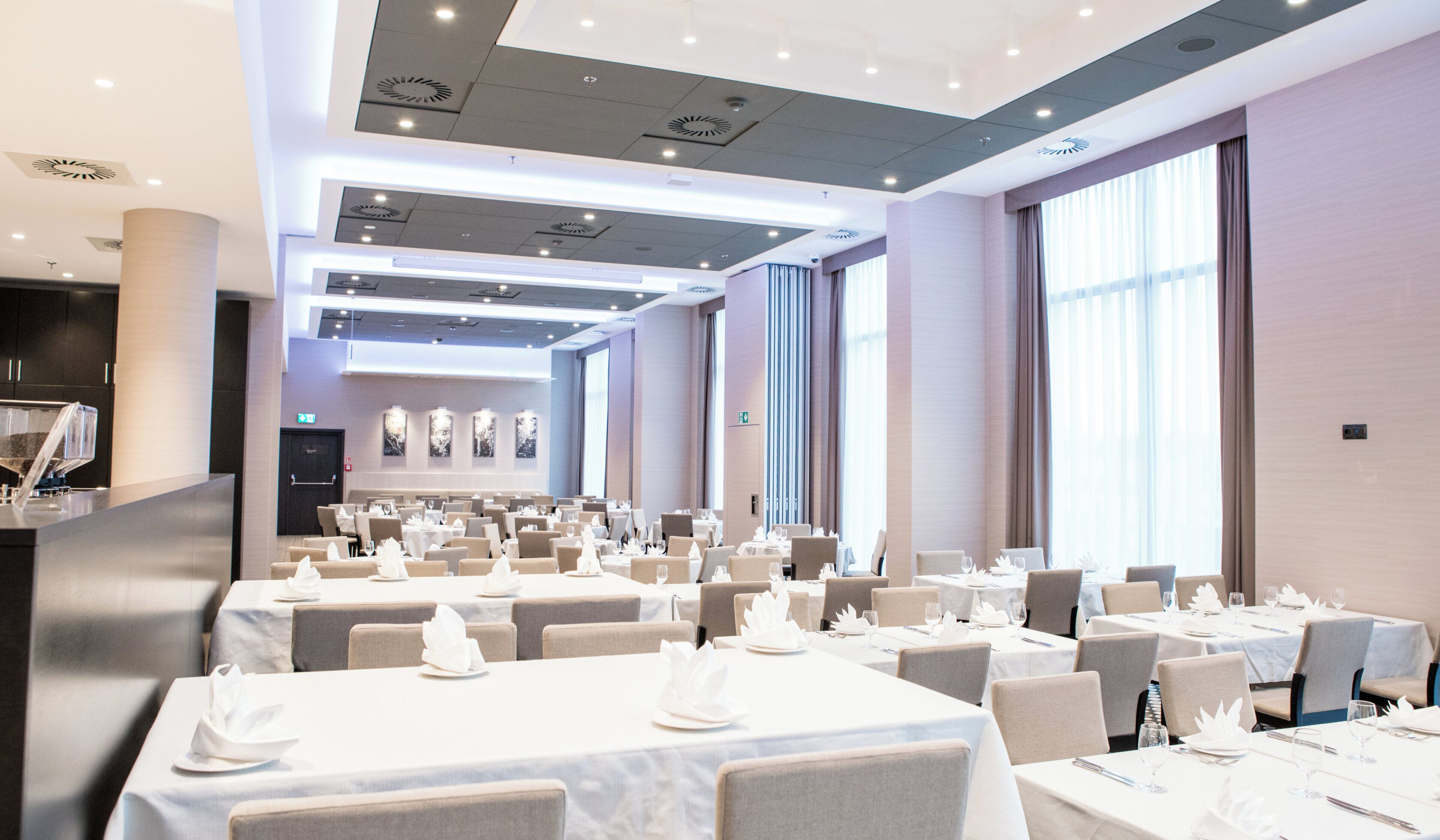 A hotel dining area with multiple tables set for guests, featuring a modern ceiling design and large windows allowing natural light.