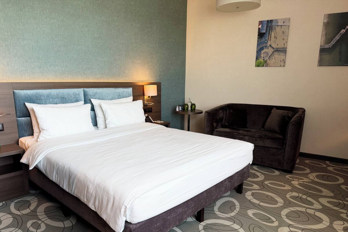A hotel room with a large bed, a dark armchair, a small table with a lamp, and a wall-mounted artwork.