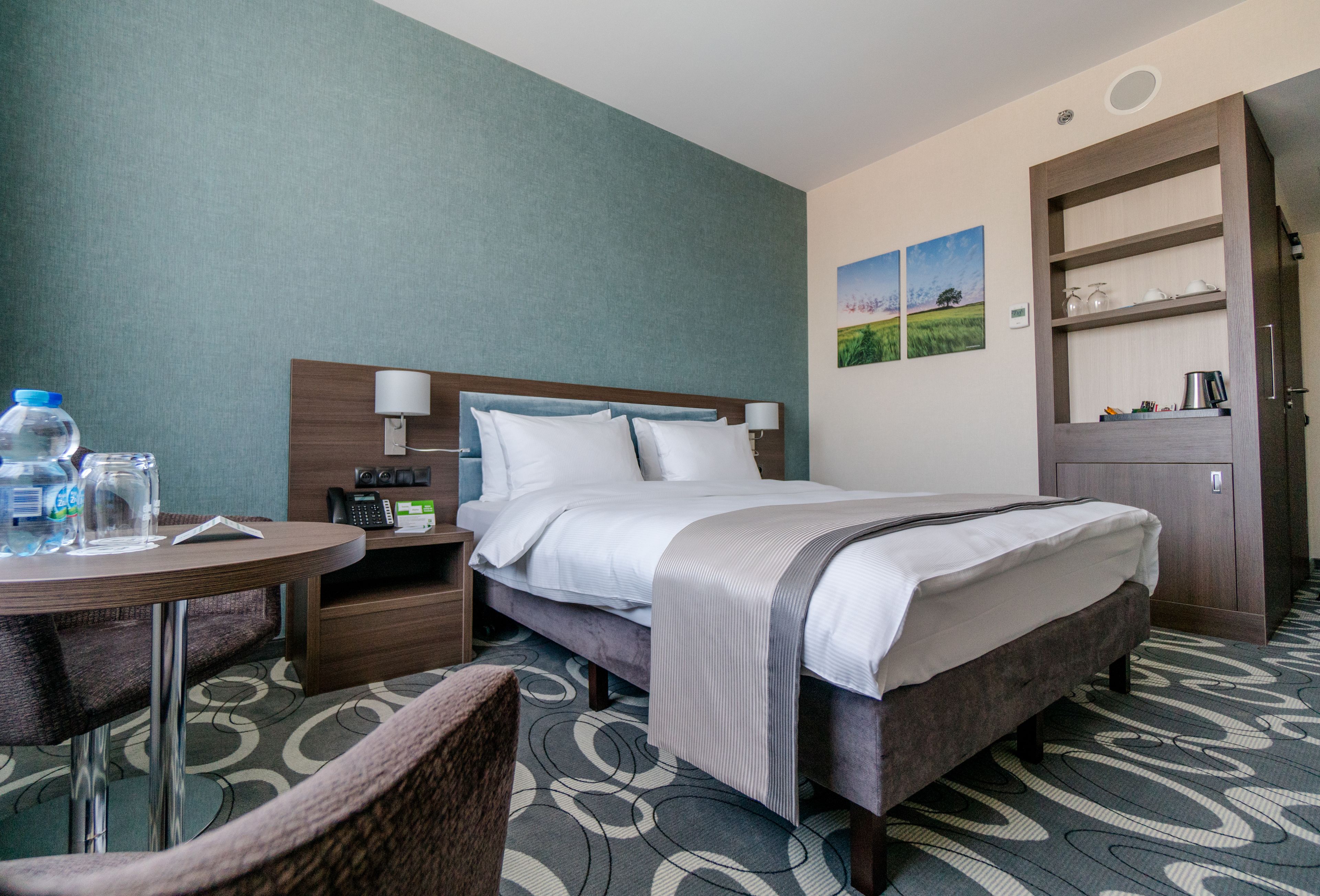 A hotel room with a bed, bedside table, lamp, and a small table with a chair.