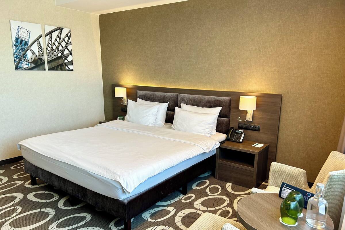 A hotel room with a bed, bedside tables, a lamp, a telephone, and a small table with a bottle and glass.