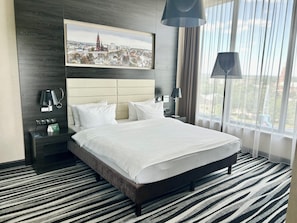 Une chambre d’hôtel avec un grand lit, une tête de lit, une table de chevet, une lampe, un tableau représentant un paysage urbain encadré et une fenêtre avec des rideaux.
