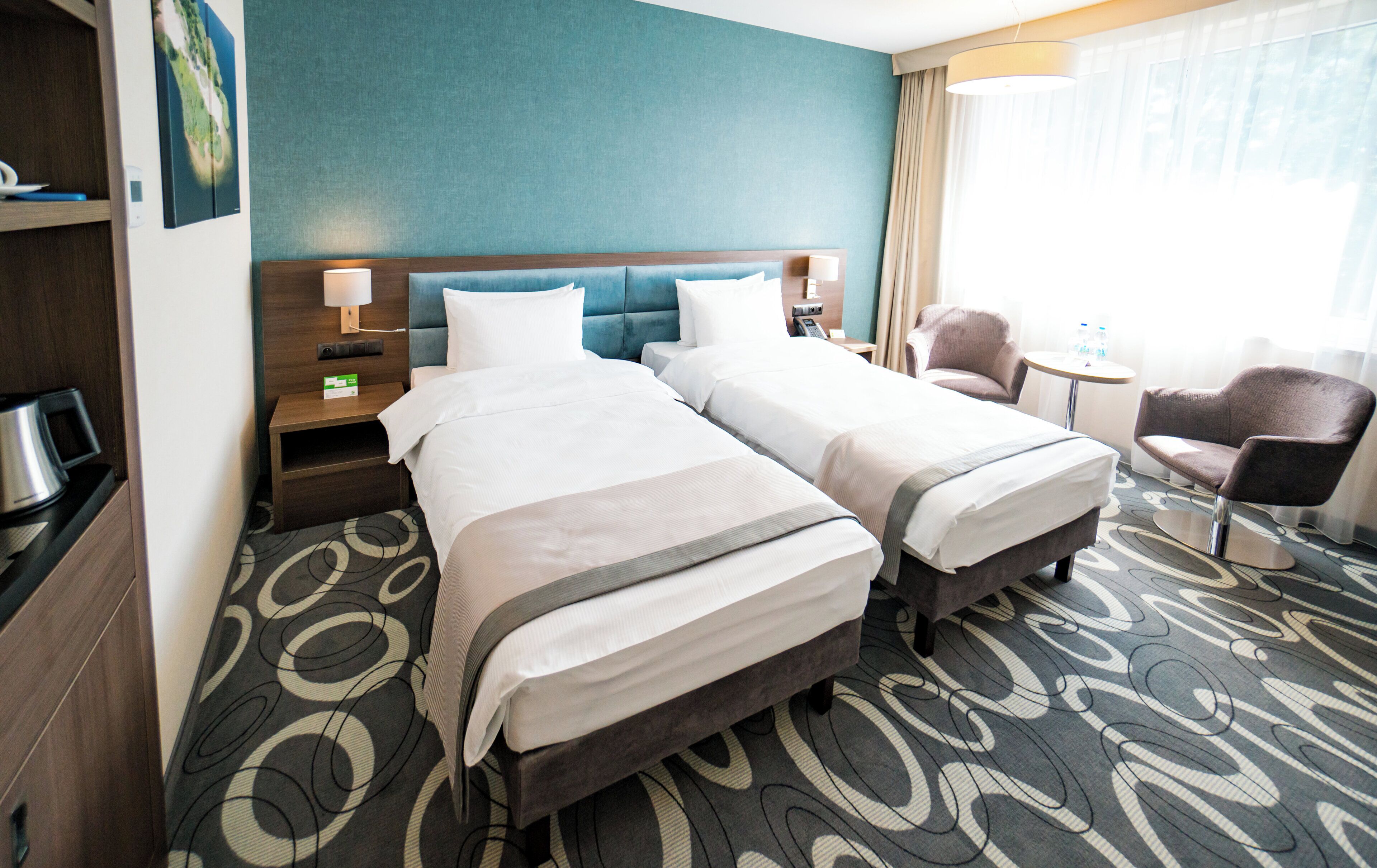 Quarto de hotel com duas camas, uma escrivaninha, uma cadeira, uma mesinha e vista para o exterior.