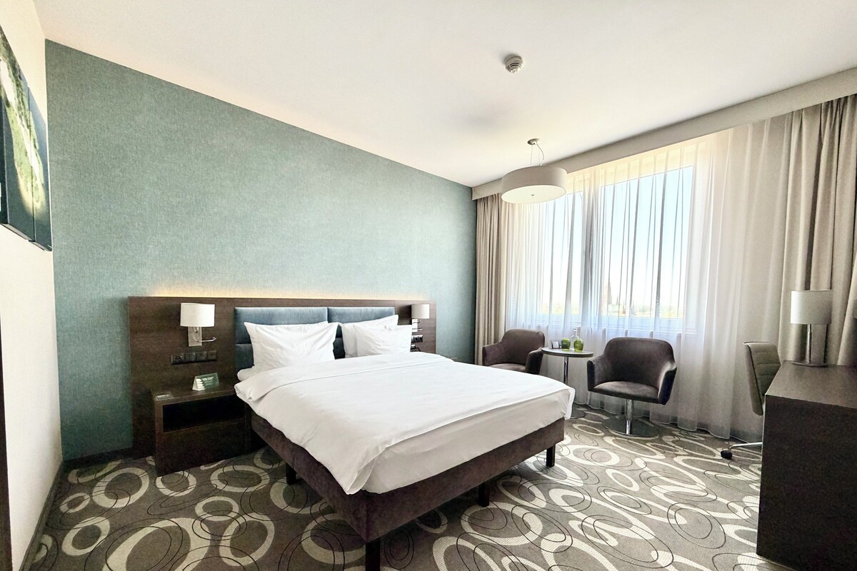 A hotel room with a bed, a desk, a chair, a lamp, and a window with curtains.