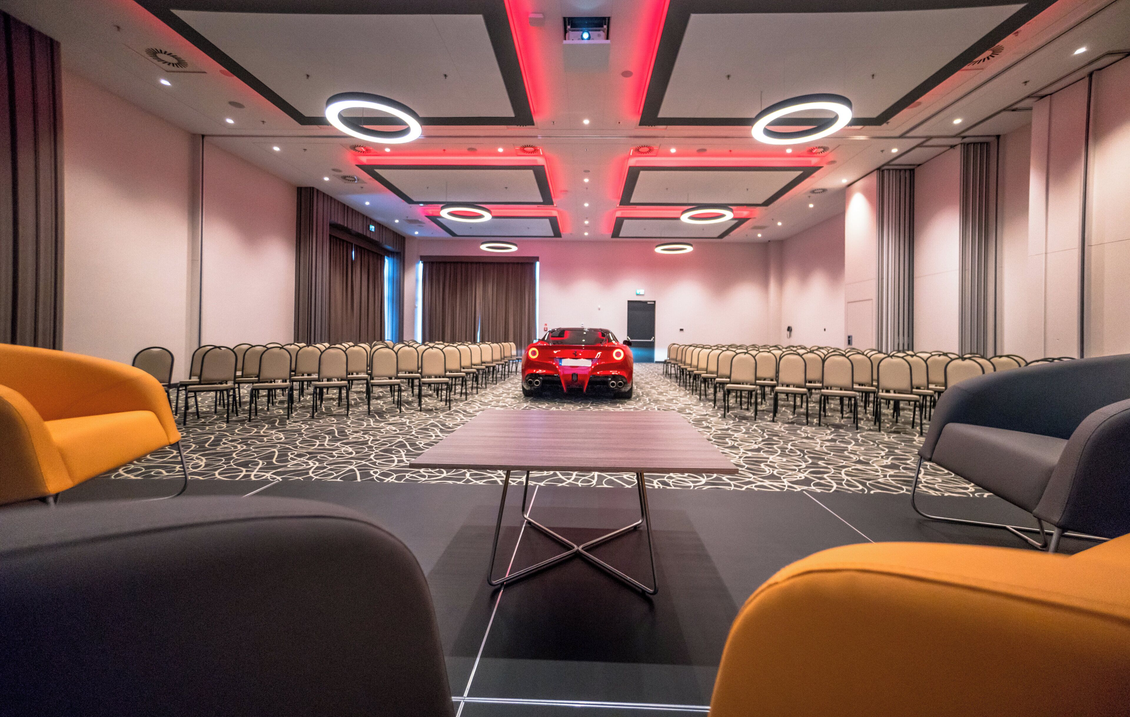 Un salón de conferencias con filas de sillas, una mesa larga y un coche deportivo rojo en el centro.