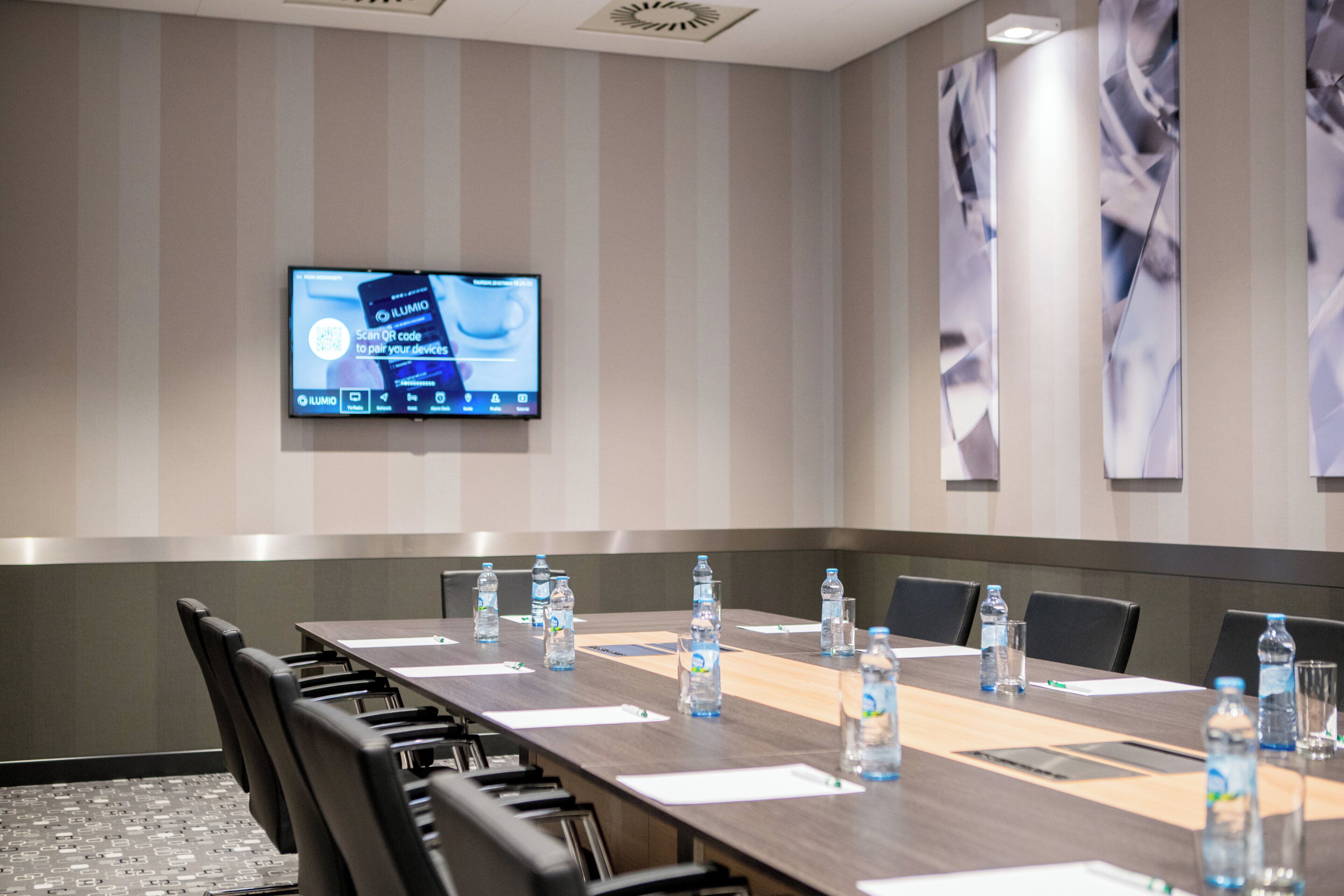 A conference room with a long table, chairs, water bottles, and a screen displaying a QR code pairing app.