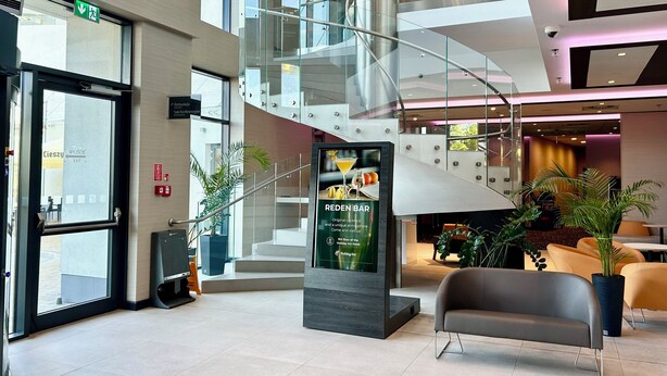 Un hall d’hôtel moderne avec un escalier en verre, un comptoir d’accueil et un panneau indiquant l’emplacement du Red Bar.