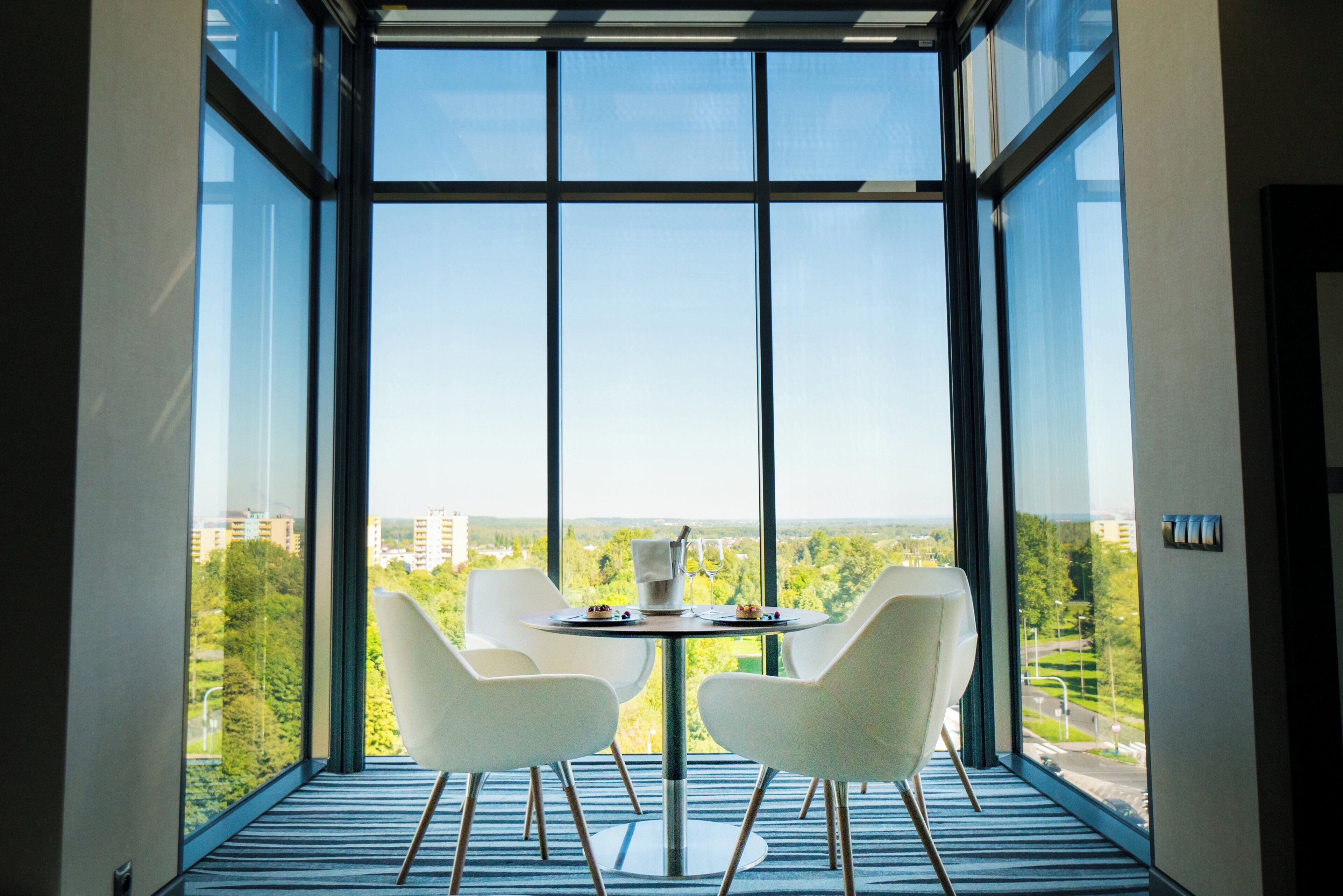 A modern room with a large window, two white chairs, a small table, and a view of a cityscape.