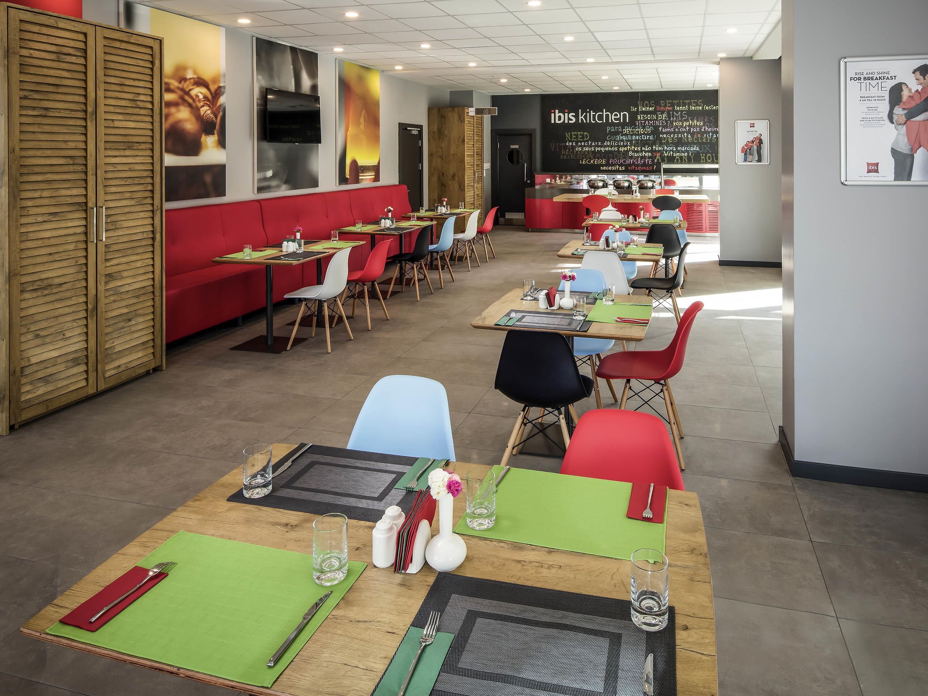 A modern restaurant with wooden tables and colorful chairs, a red booth seating area, and a kitchen counter in the background.
